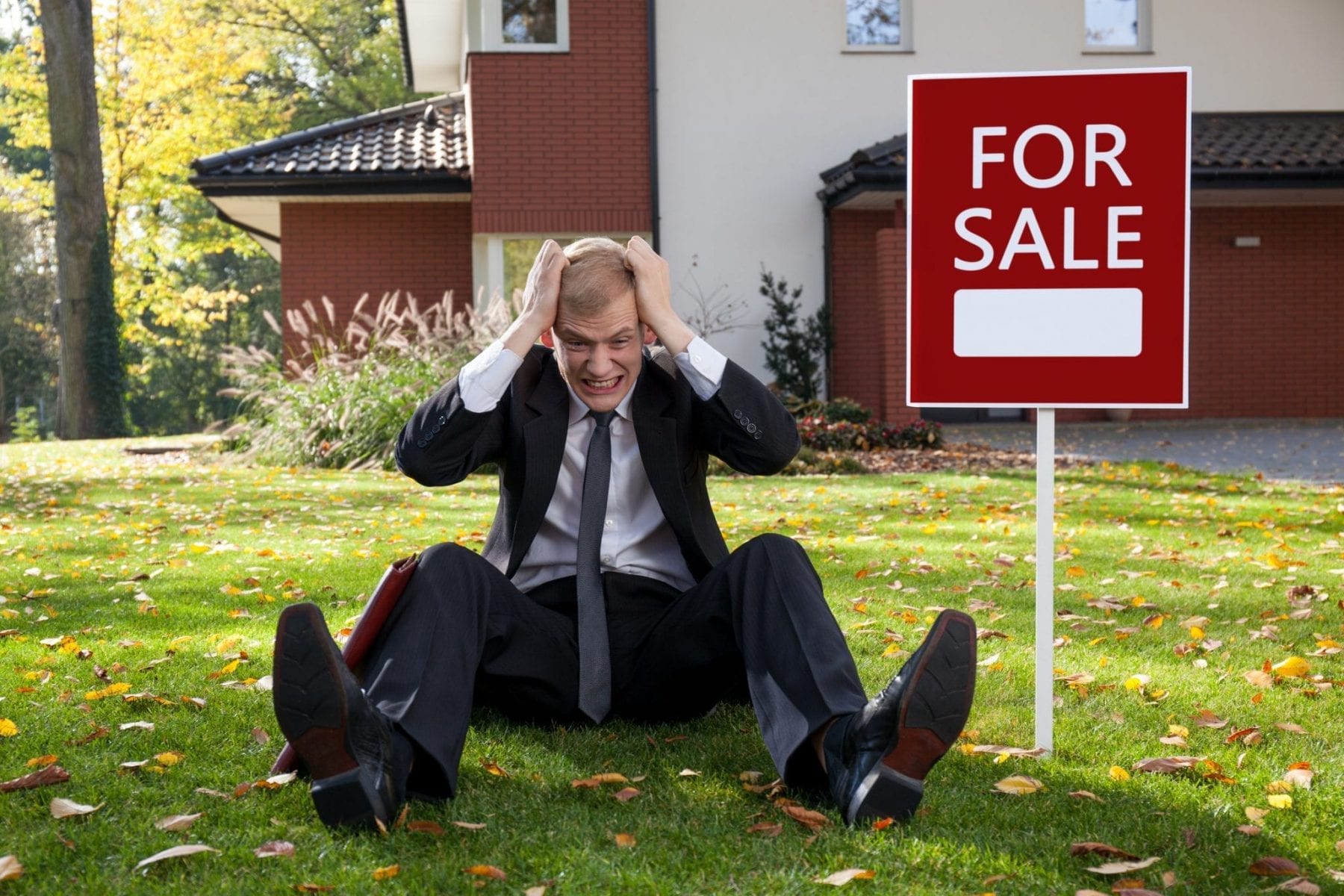 Man in suit sitting next to For Sale sign pulling on his hair