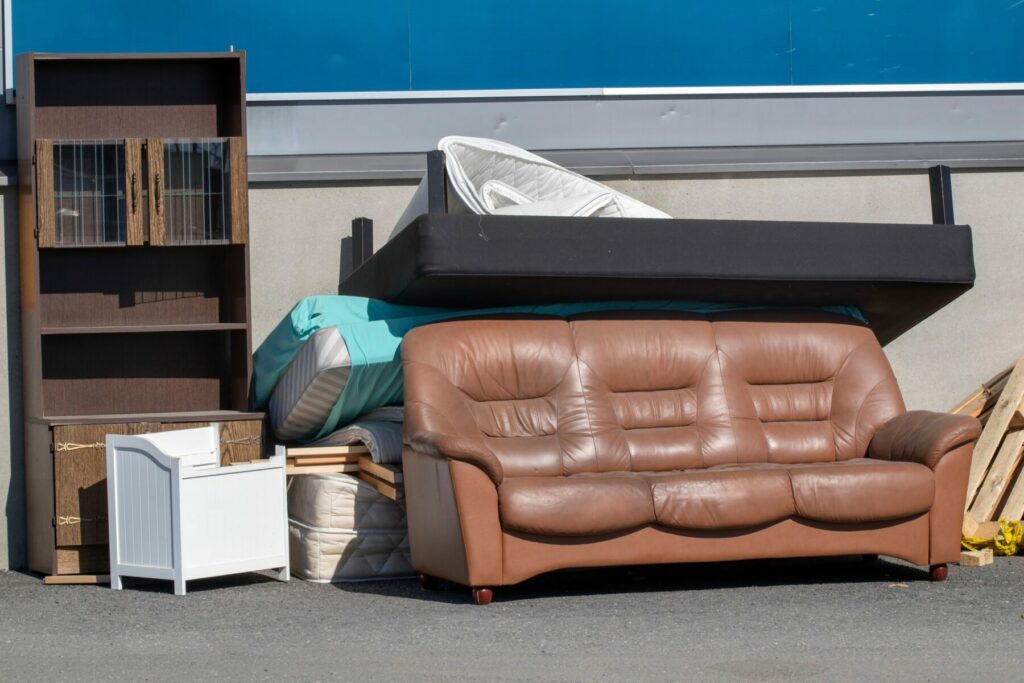 A pile of used furniture outdoors with a couch, a cabinet and a bedframe among other items