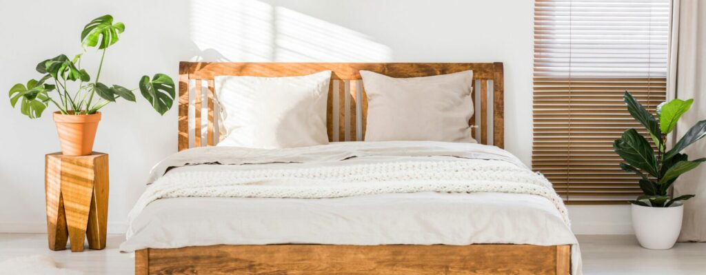 Wooden bed with bedding, pillows and blanket against white wall in a bright sunny bedroom interior