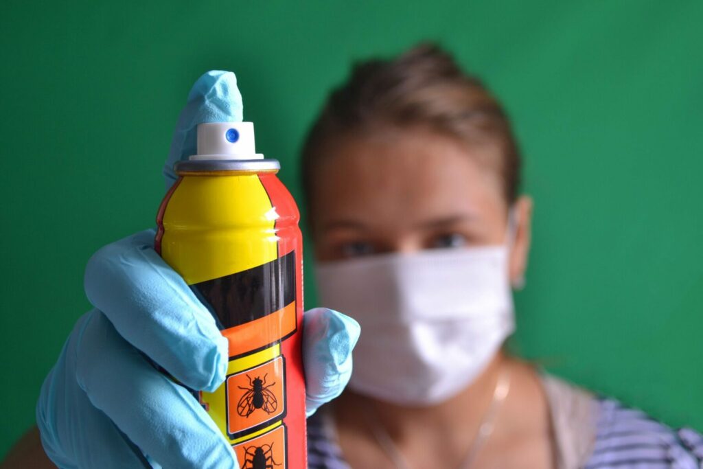 a woman wearing blue nitrile gloves and a face mask holds up an aerosol bug spray can