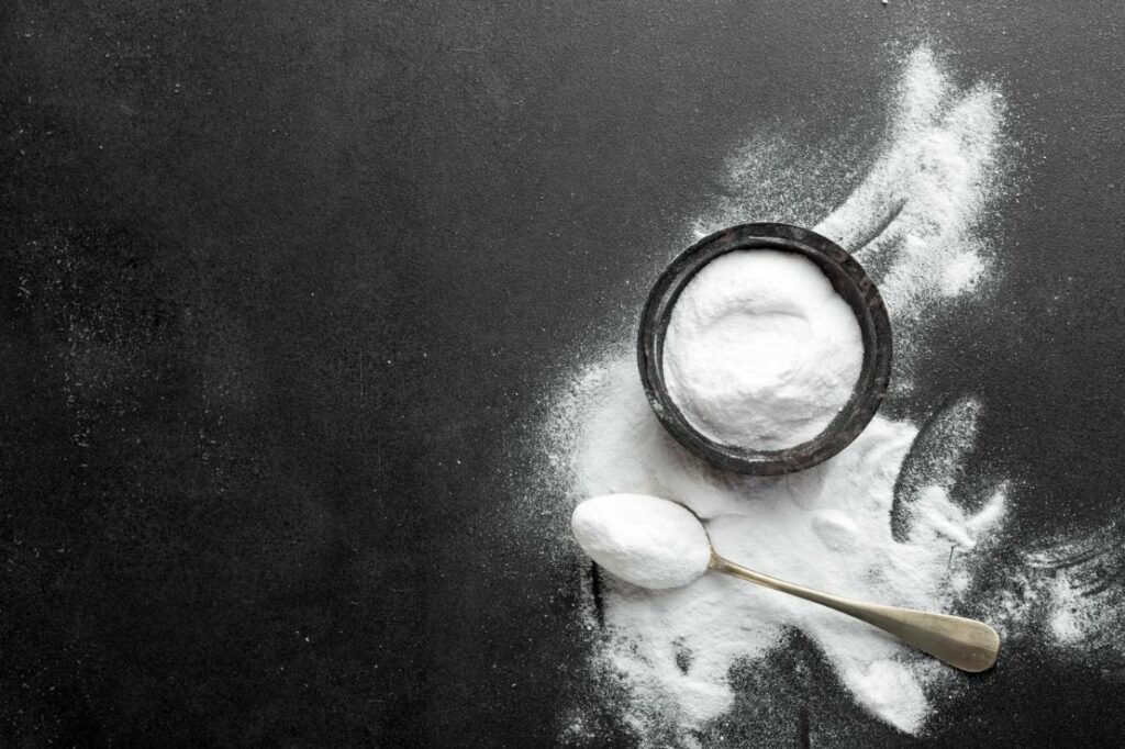 A small black bowl of baking soda that has also been spilled sits on a black counter with a spoon full of baking soda sitting beside it