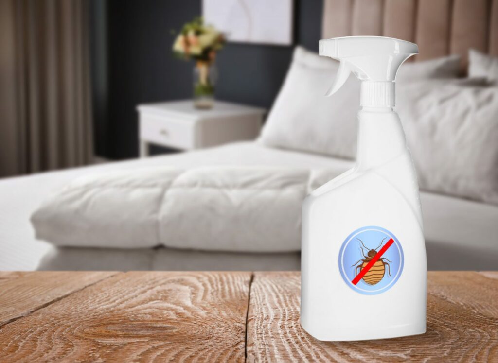 a white unlabeled spray bottle with a sticker showing an X over a bed bug sits on a table in front of a bed