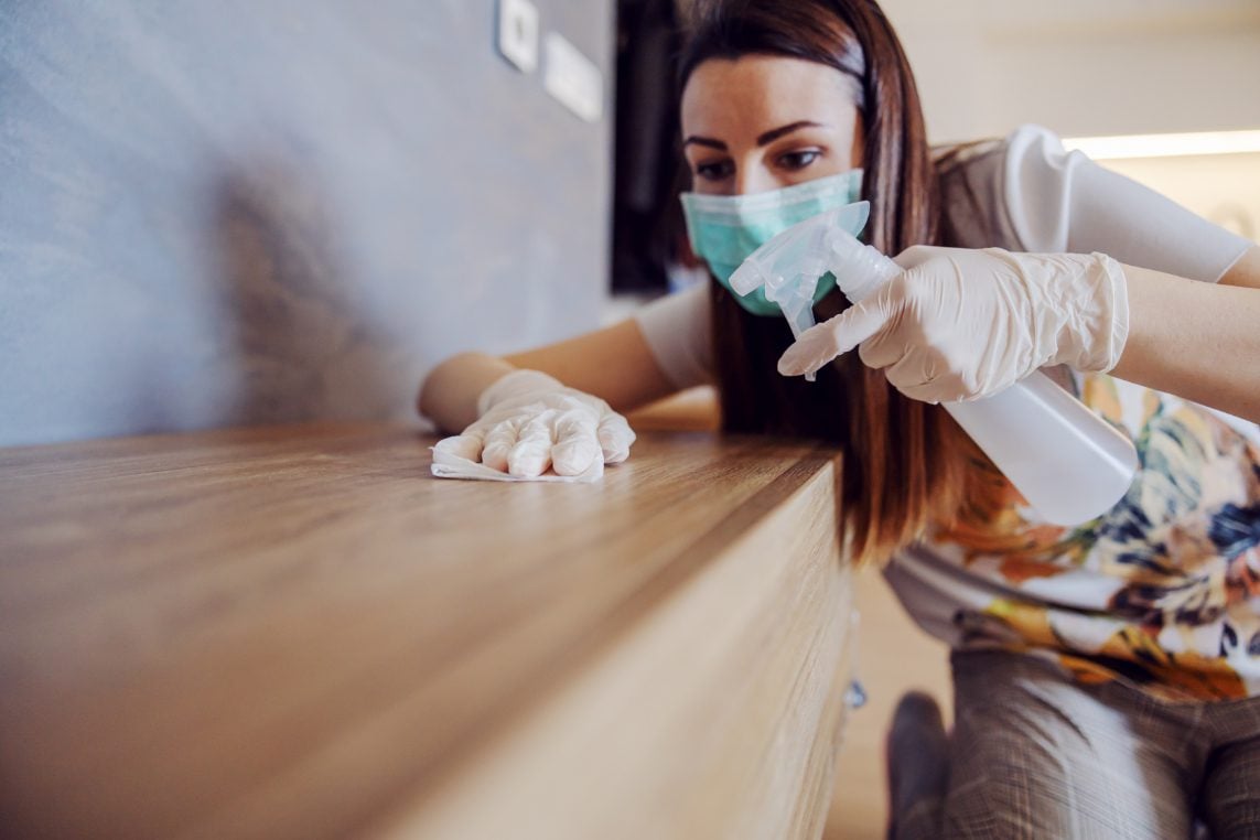 A woman wearing a blue medical face mask and white gloves uses a spray bottle to clean a wooden table