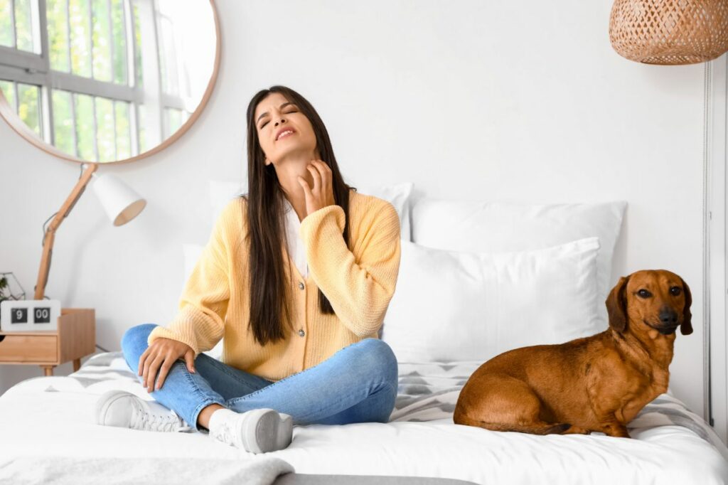 A woman in a yellow sweater and jeans sits in her bed, scratching her neck while her small brown dog sits beside her