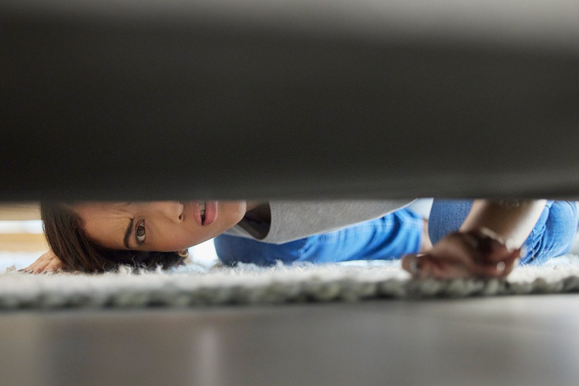 A person on their hands and knees looks underneath a couch